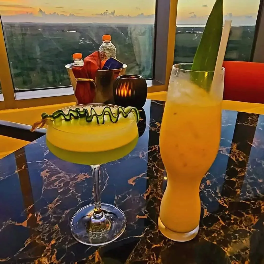 Dos cócteles amarillos con decoraciones verdes sobre una mesa junto a una ventana al atardecer.