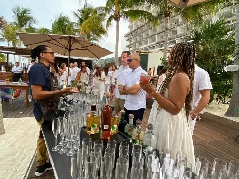 Una persona con mandil conversa con invitados junto a una mesa con copas y botellas al aire libre.