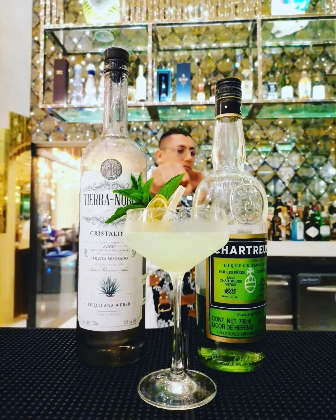 Cóctel claro en copa con una decoración de cítrico y hoja verde, junto a botellas de tequila y licor de hierbas.