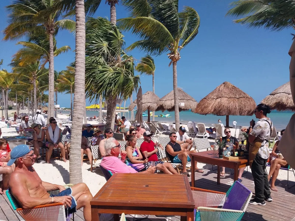 Grupo reunido para una presentación de bebidas junto a una playa con palmeras y sombrillas de palma.