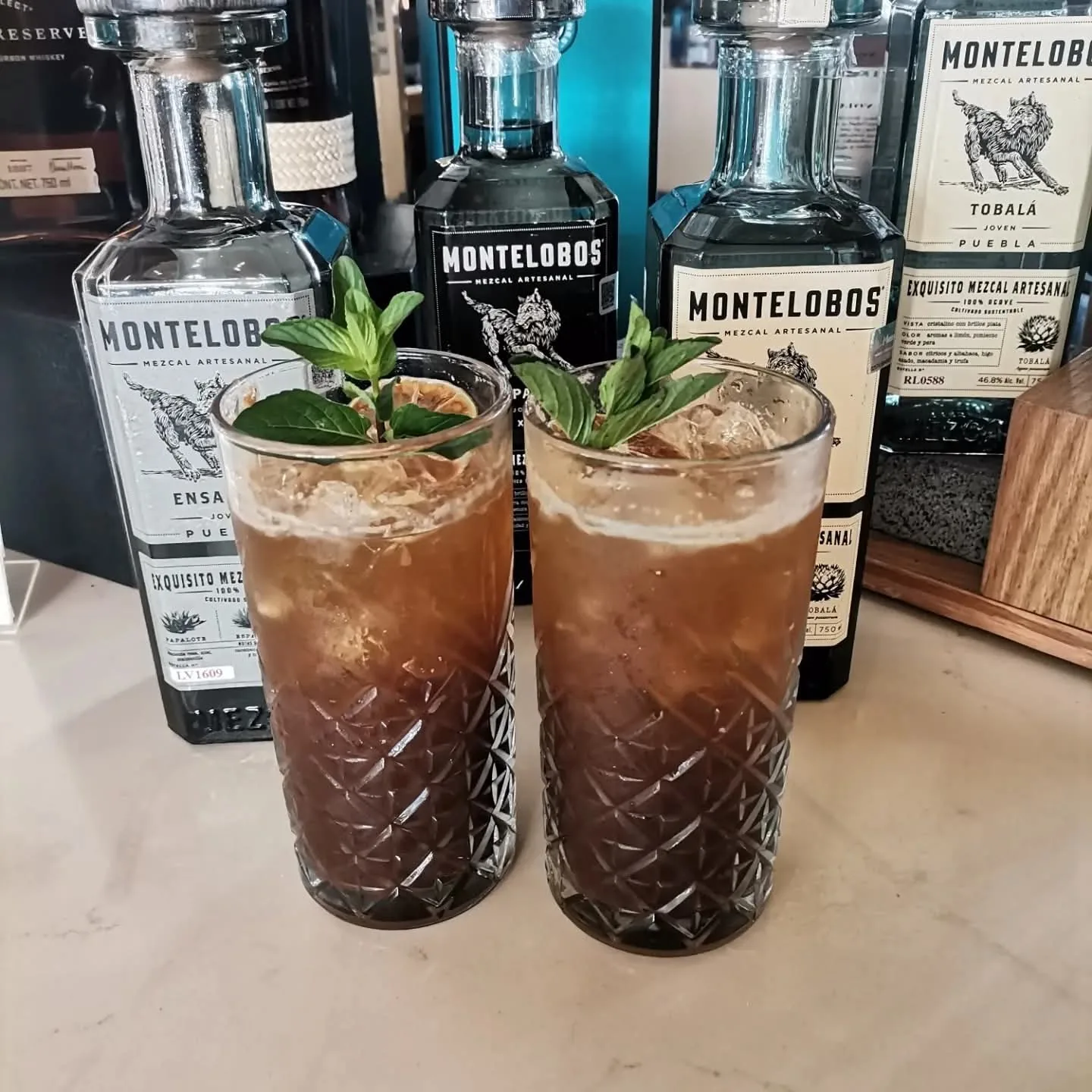 Dos cócteles oscuros con hielo y hojas verdes delante de botellas de mezcal.