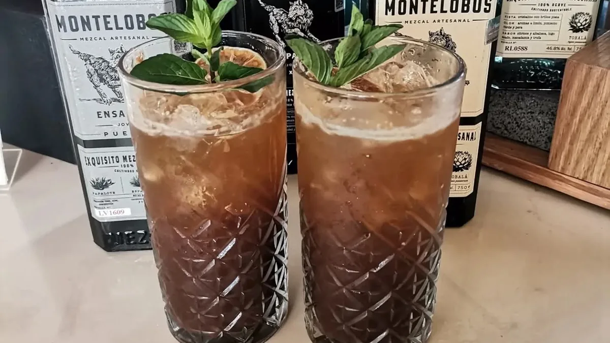 Two dark cocktails with ice and green leaves in front of mezcal bottles.