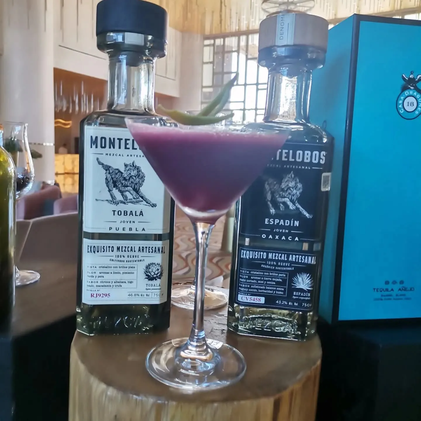 Cóctel morado en copa con una hoja verde, junto a botellas de mezcal.