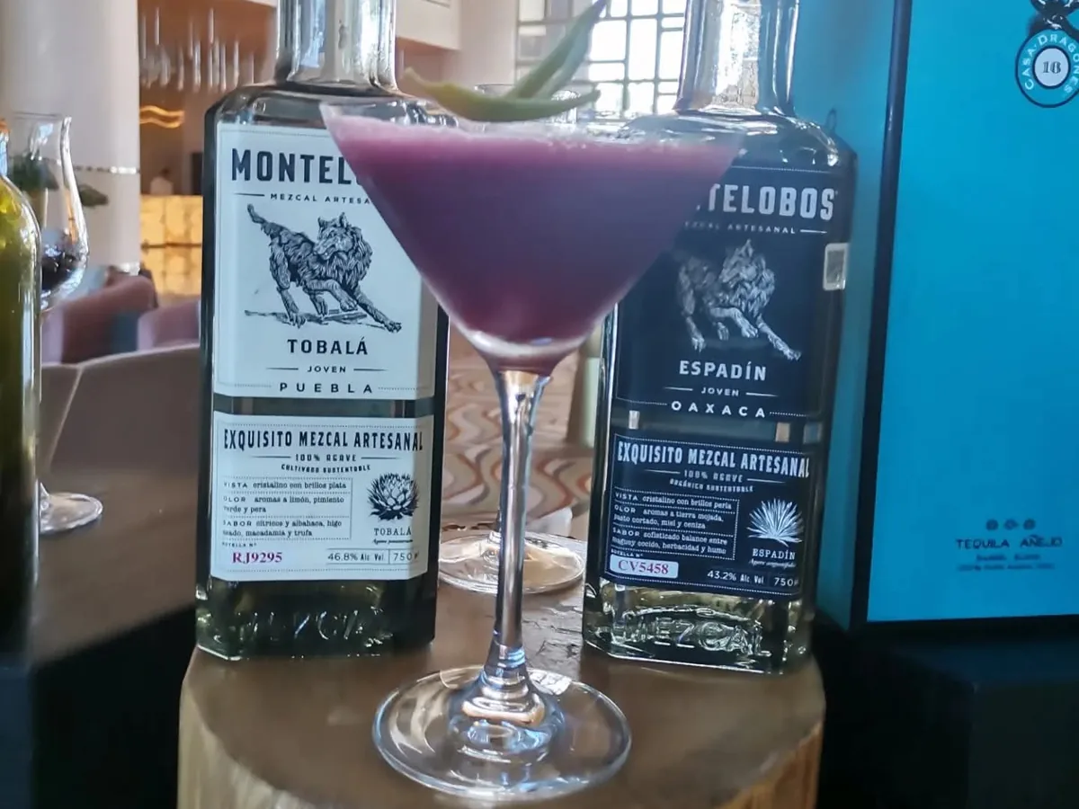 Cóctel morado en copa con una hoja verde, junto a botellas de mezcal.