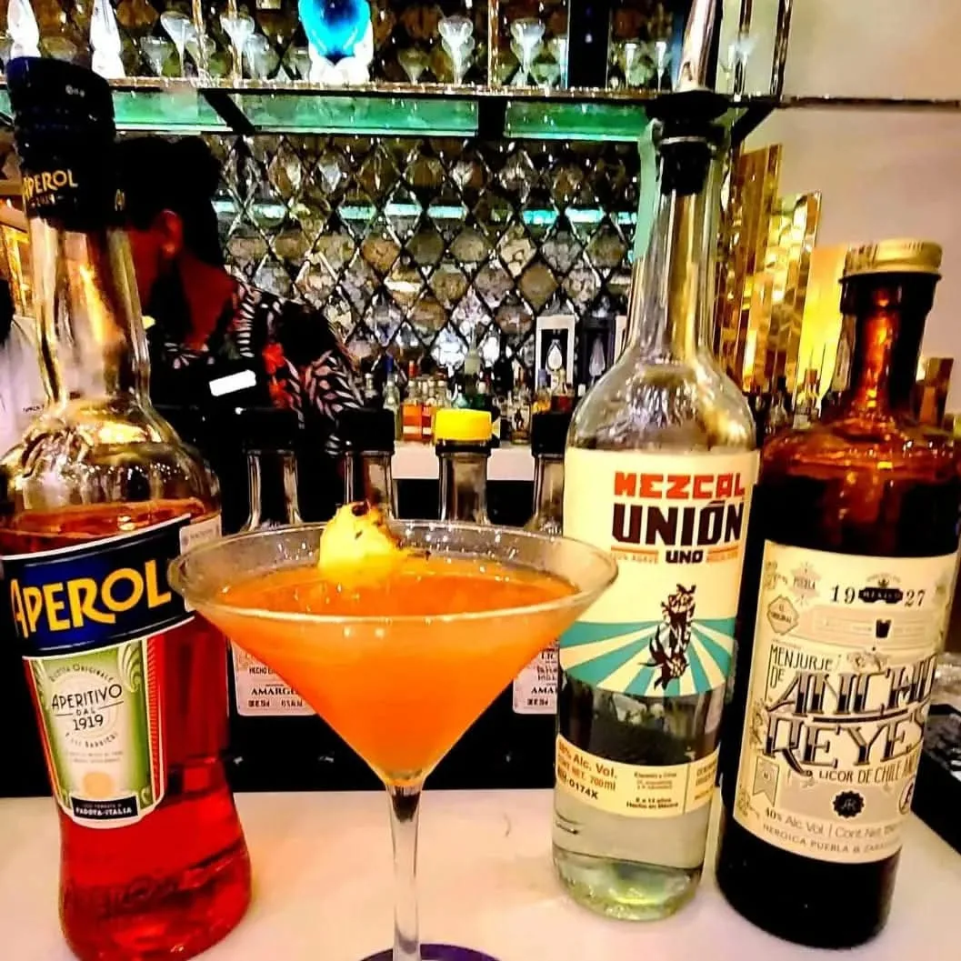 Cóctel naranja en copa junto a botellas de aperitivo, mezcal y vermut sobre una barra.