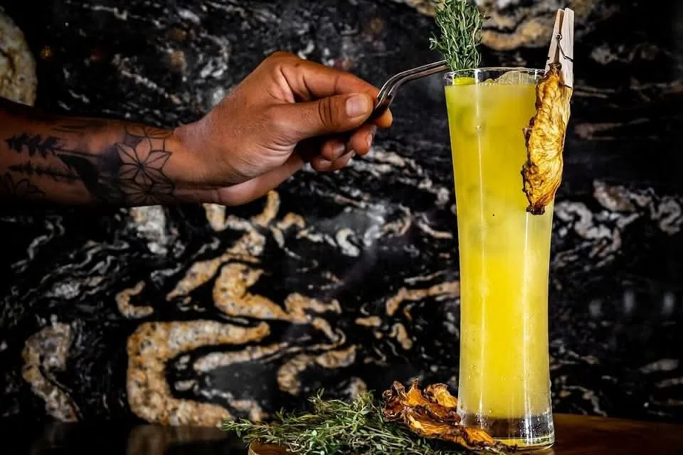 Una mano con tatuajes decora un cóctel amarillo en vaso alto con una pieza de piña dorada.