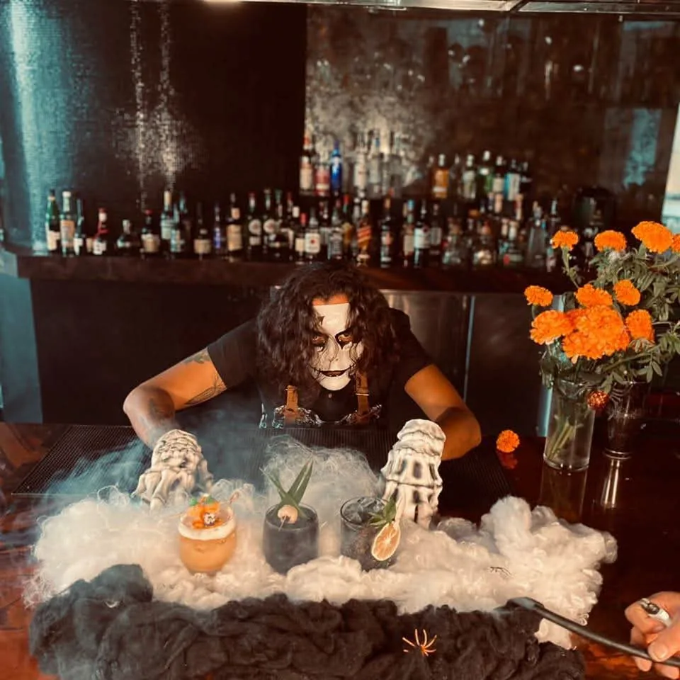 Persona con maquillaje de calavera presenta bebidas entre niebla y flores de cempasúchil en una barra.